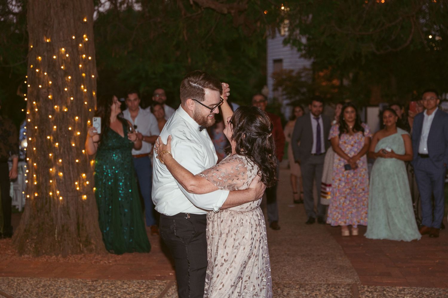 Wedding guests watch as a couple shares a romantic dance moment under tree-strung lights at dusk.
