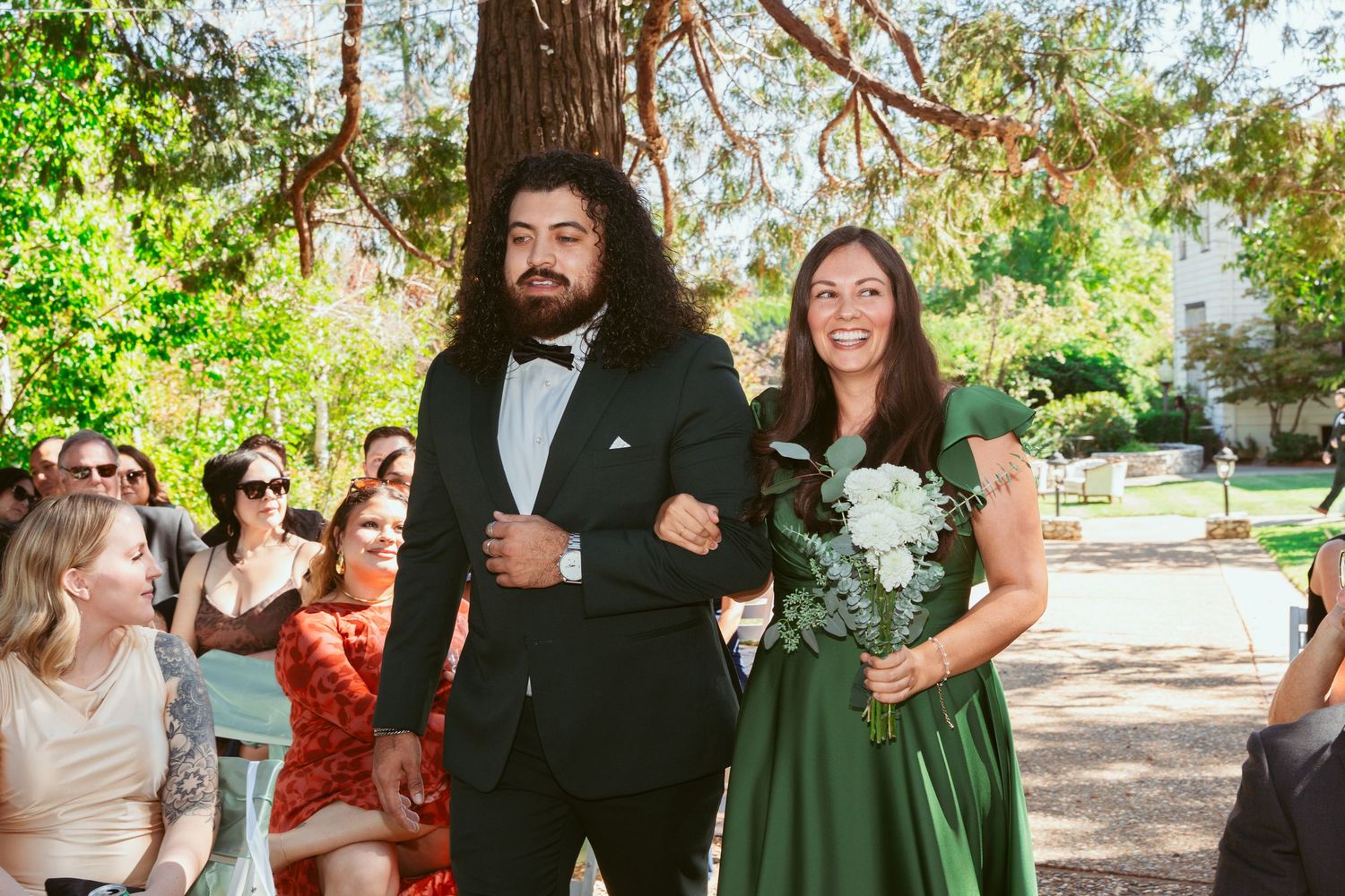 Wedding guests gather outdoors under trees for a casual celebration in a natural garden setting.