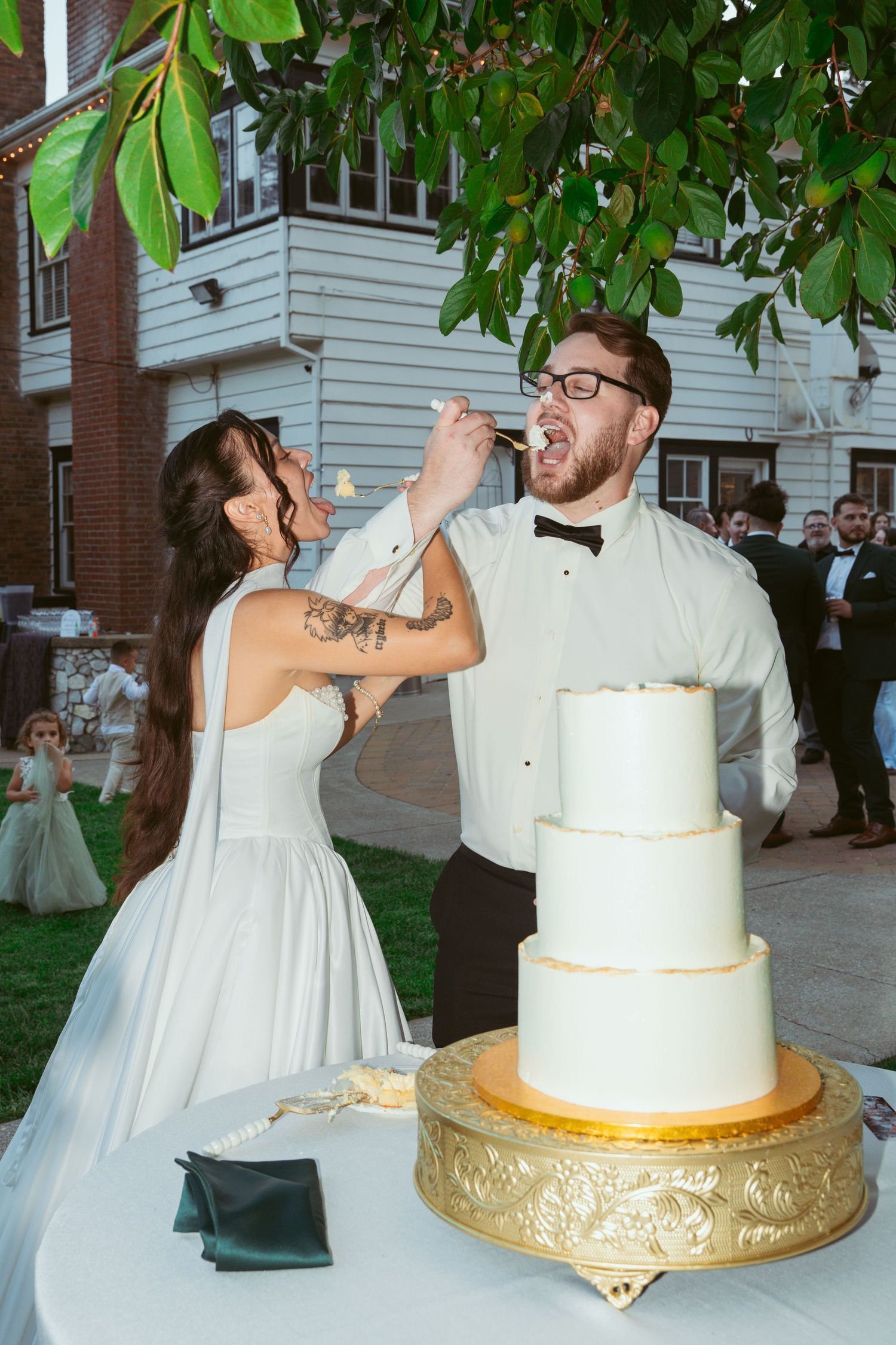 Wedding couple sharing cake-cutting moments outdoors under tree branches with white tiered cake.