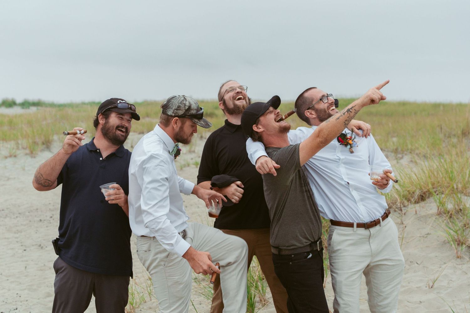 A group of groomsmen strike fun poses while celebrating on a sandy beach.