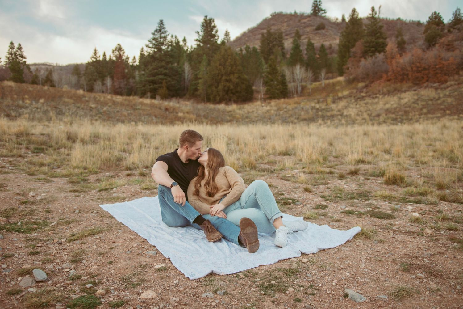 Couple relaxing on a light blue blanket in a grassy mountain meadow during golden hour.