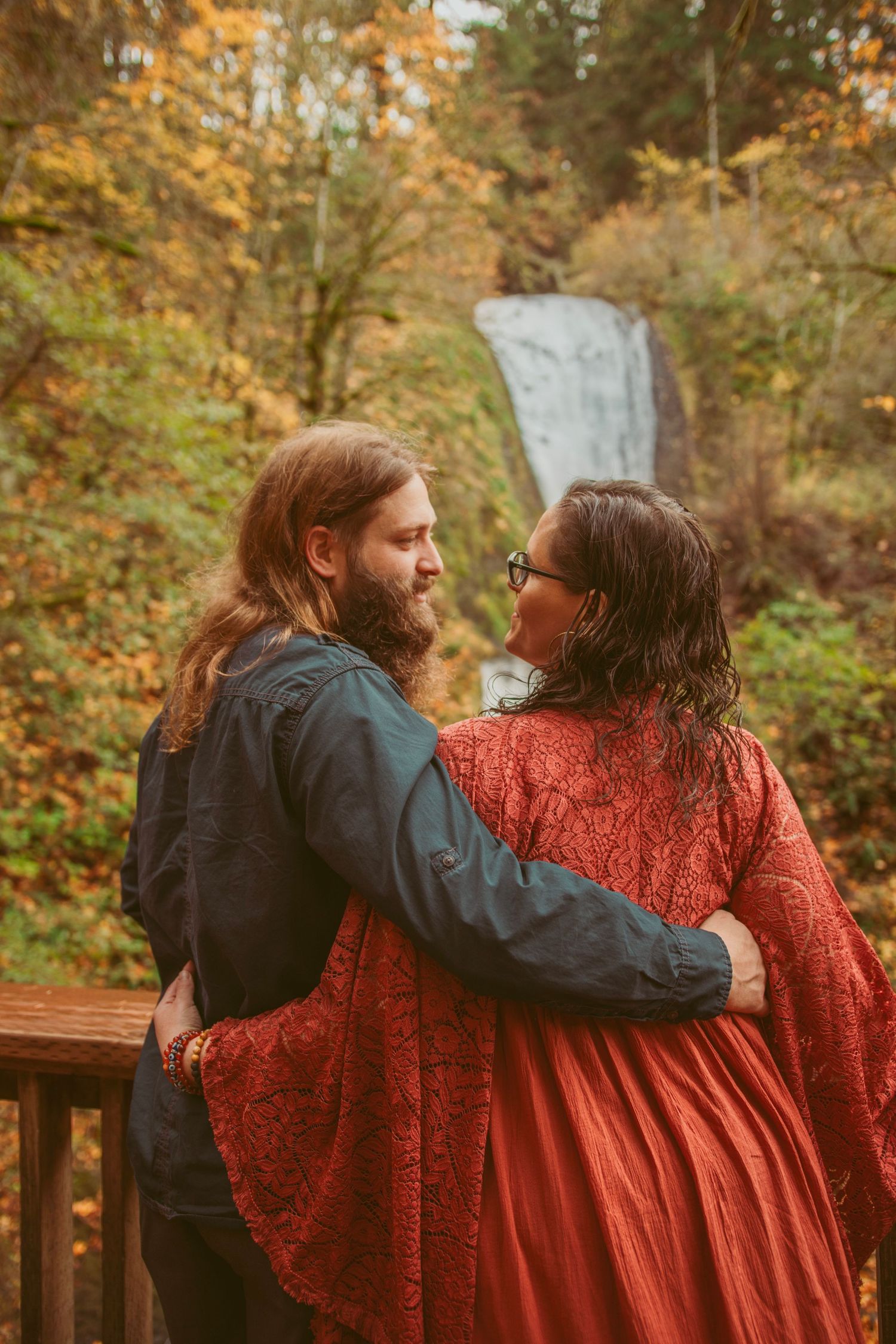 A couple in red and blue clothing embracing on a wooden deck during autumn.