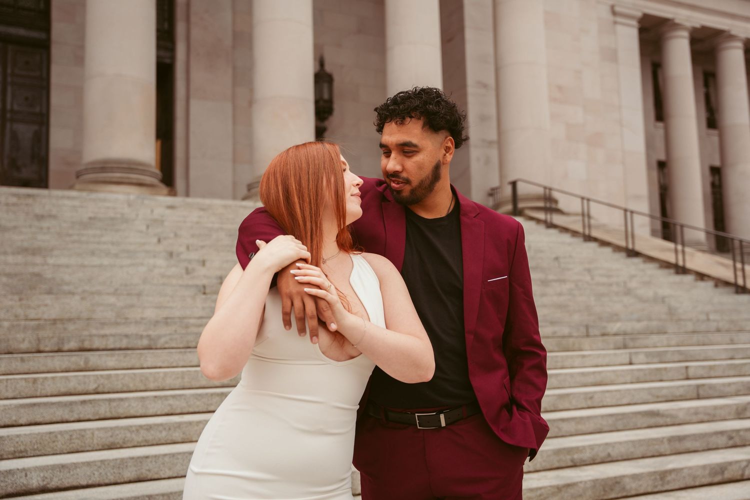 Dancing couple in burgundy and white outfits pose on stone steps of a historic building.