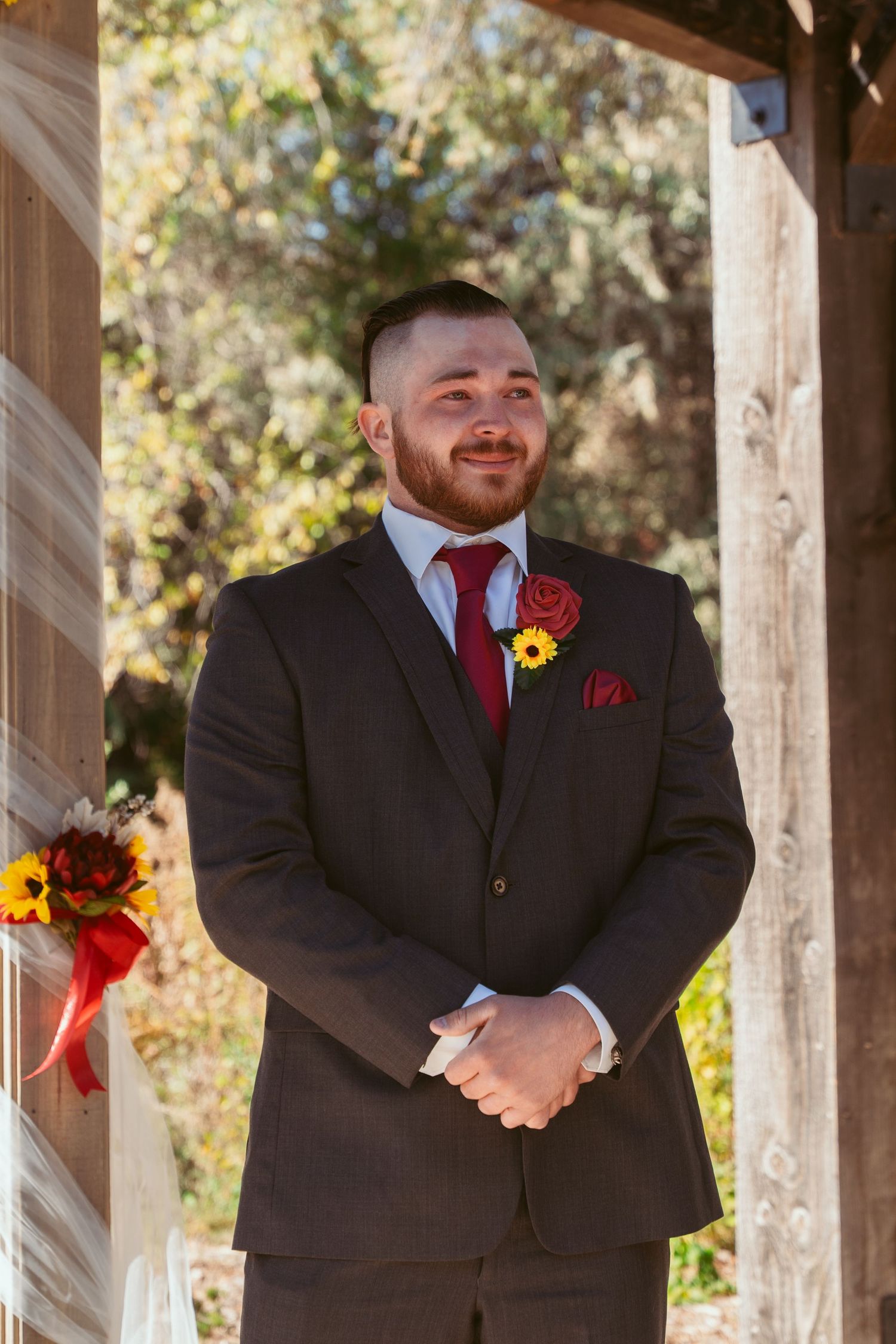 Attendant in dark suit with red boutonniere stands at altar during outdoor ceremony.