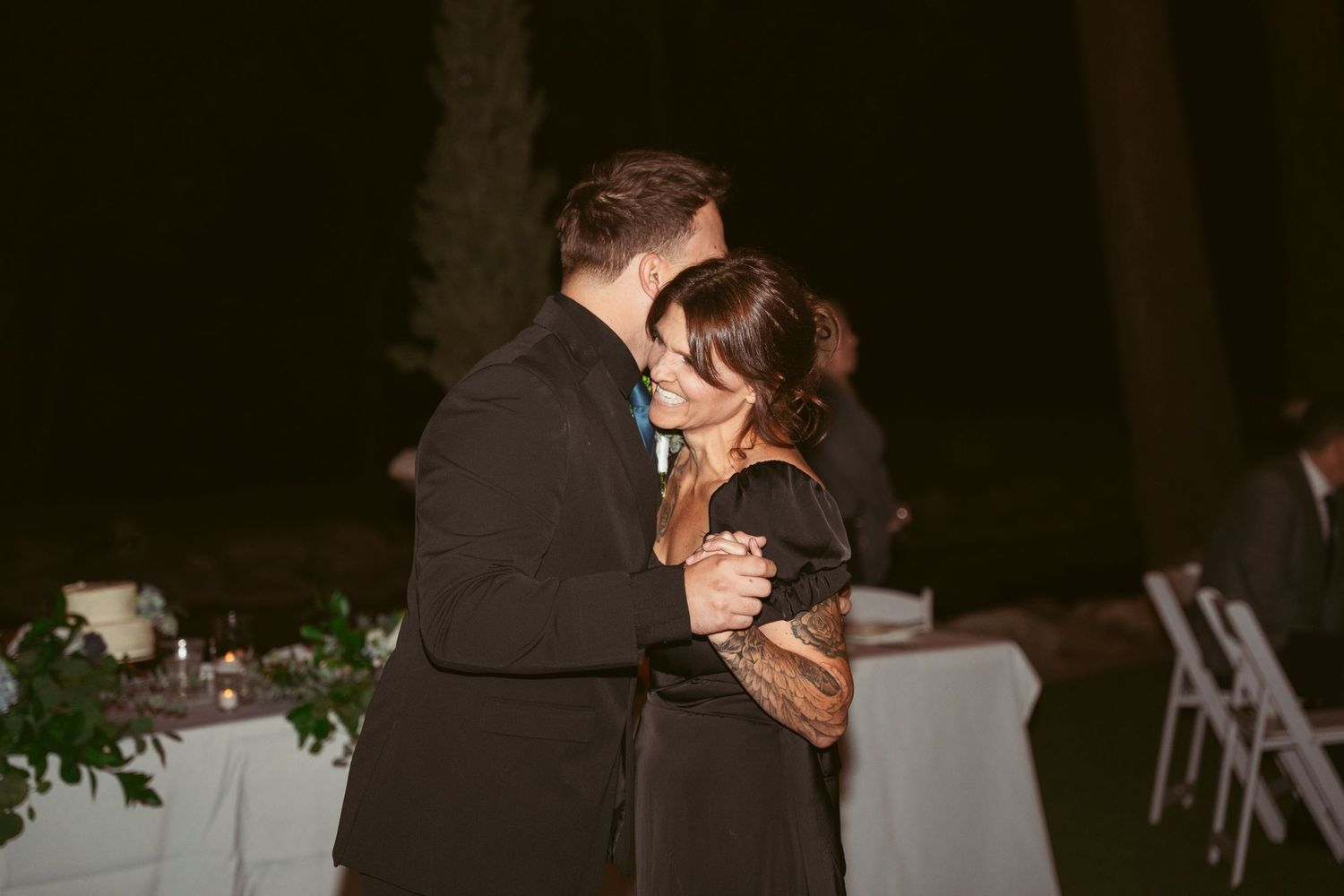 Wedding reception guests slow dancing under romantic outdoor lighting at dusk.
