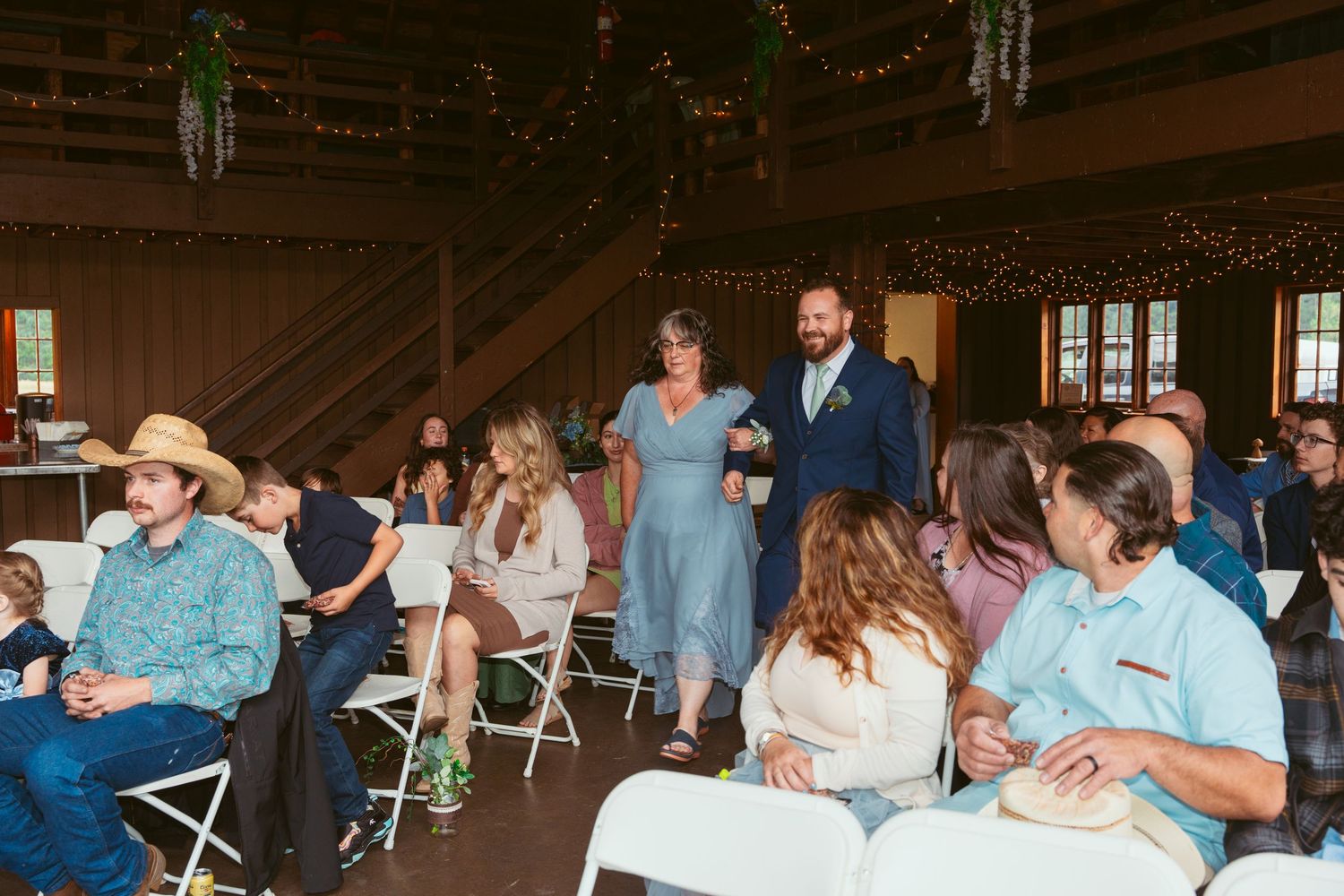 Wedding guests seated in folding chairs watching ceremony in rustic barn setting.