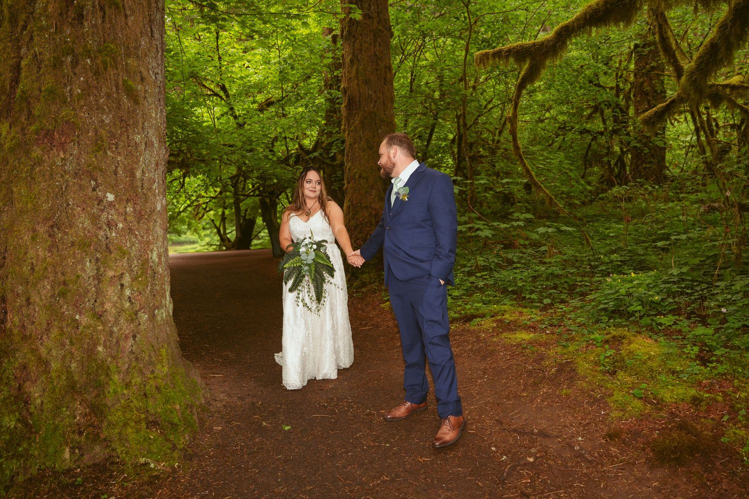 A bride in white and groom in navy walk through a serene wooded area.