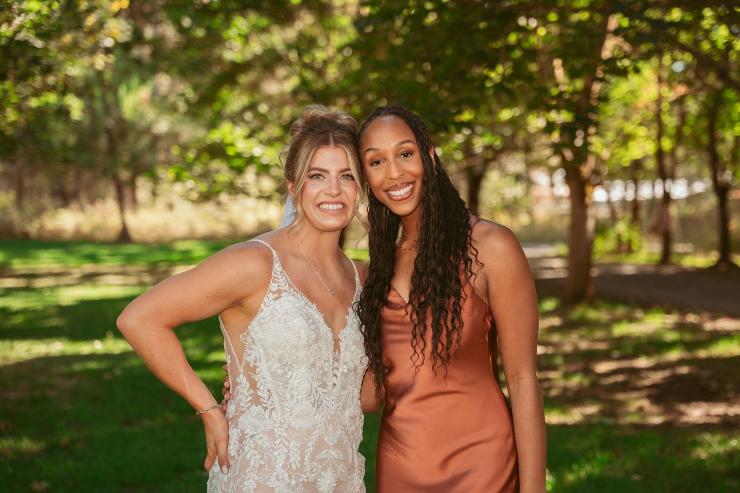 Two friends in formal dresses smile together in a sunlit garden setting.