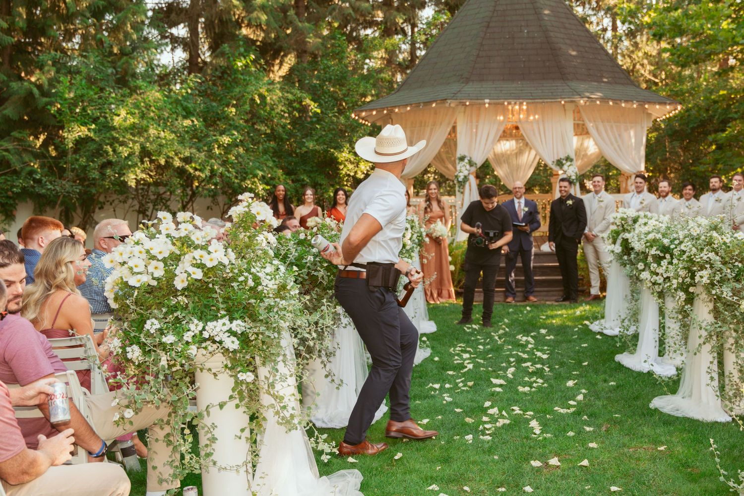 Garden wedding ceremony site with white floral arrangements and scattered petals on grass.