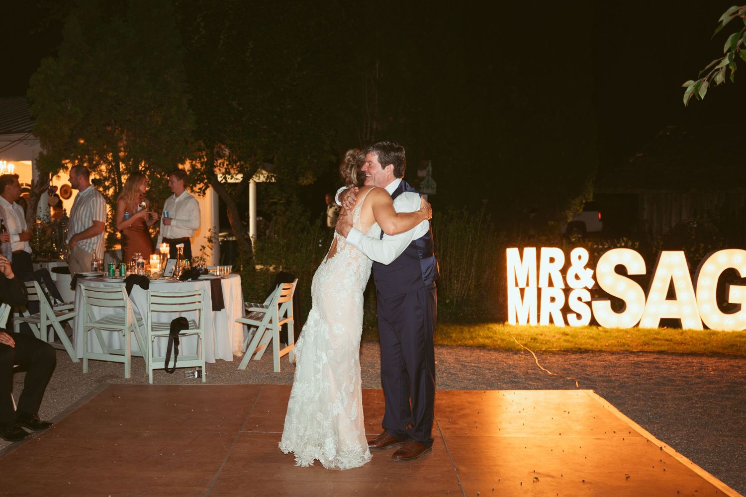 Couple shares their first dance near illuminated 'MR & MRS' sign at outdoor wedding reception.