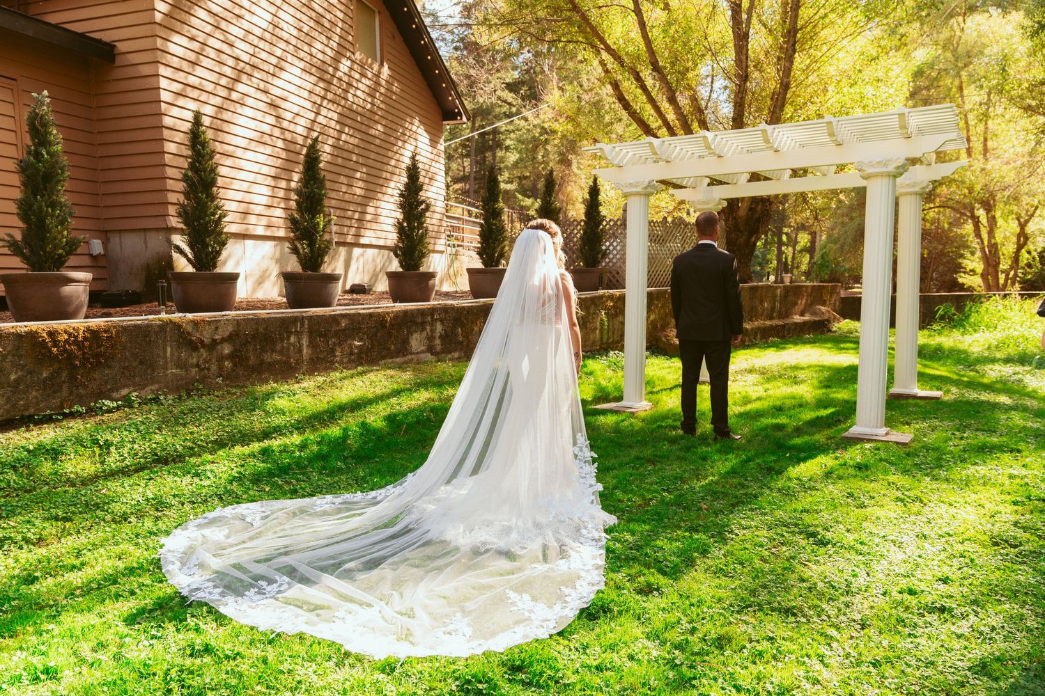 A flowing cathedral-length veil trails behind on a lush green lawn during an outdoor wedding ceremony.