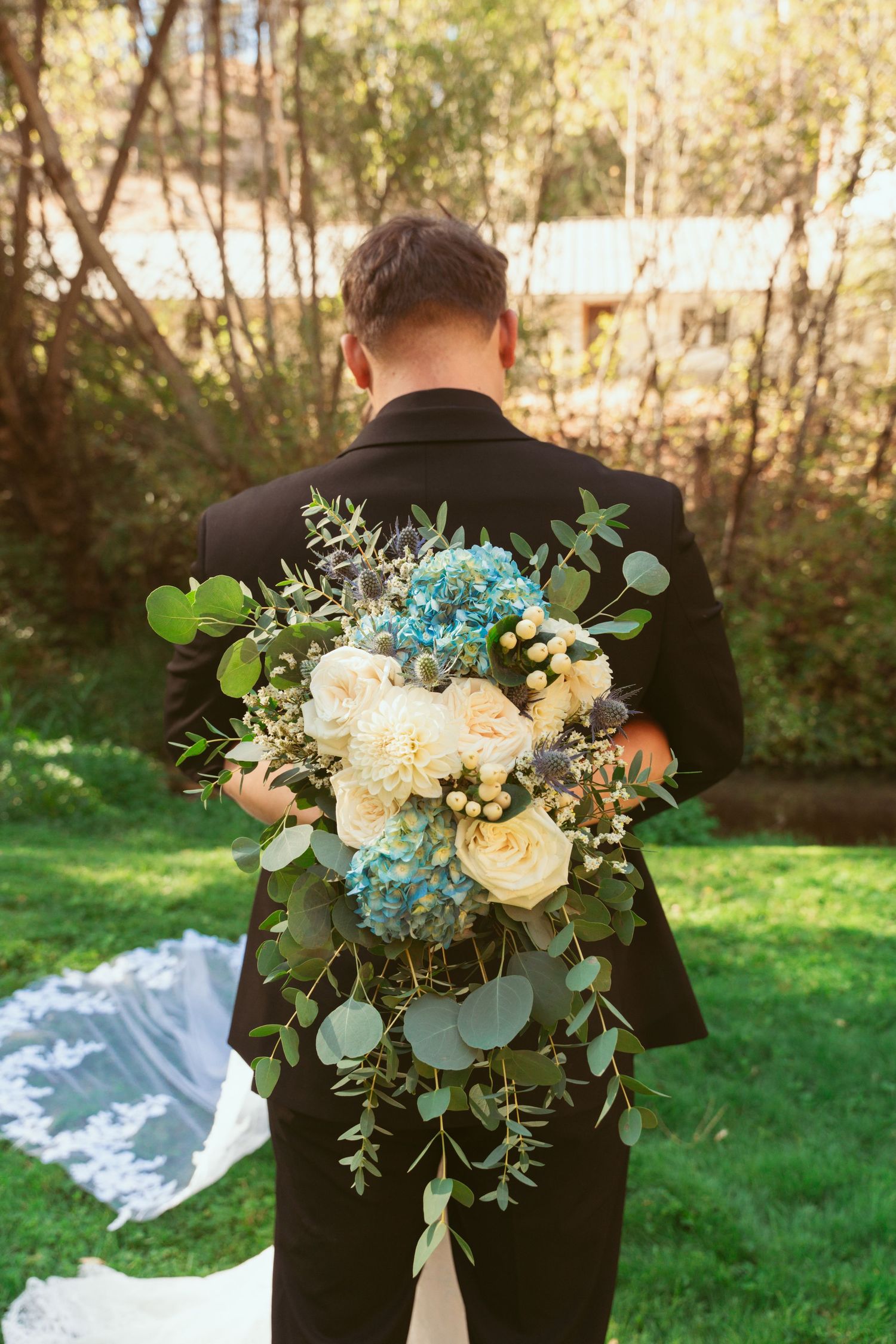 Wedding bouquet of white roses and blue flowers with eucalyptus greenery held against a black suit jacket outdoors.