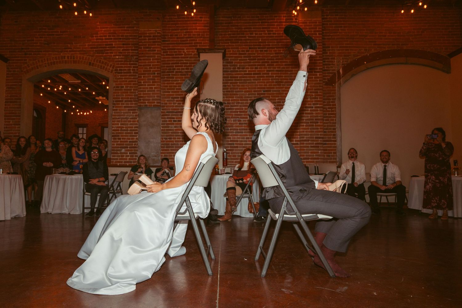 Dancers perform a synchronized move at a wedding reception in a brick-walled venue with twinkling lights.