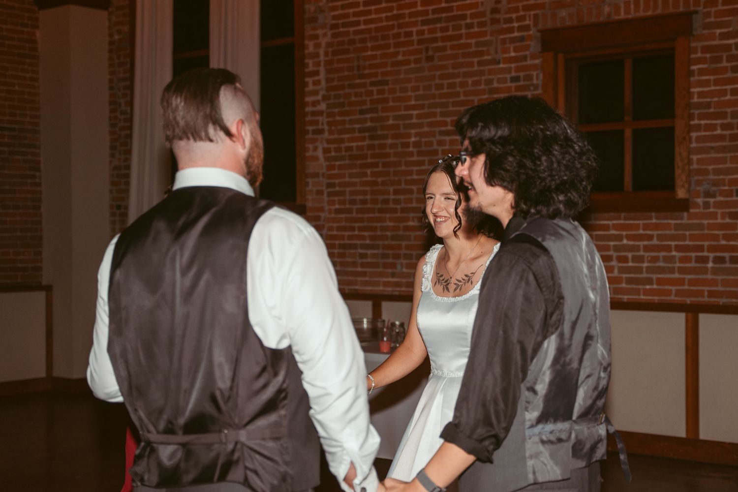 Wedding party members dance together in front of exposed brick wall with string lights.