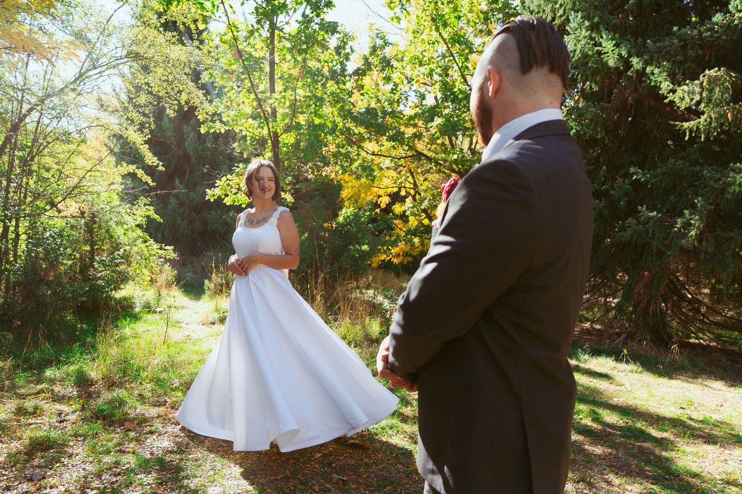 A bride in a flowing white dress twirls in a sunlit forest setting while her partner watches.