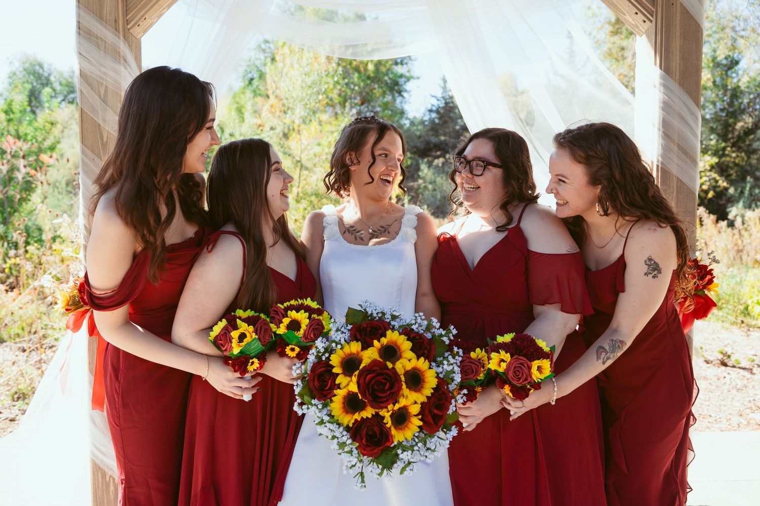 Wedding party in burgundy attire poses with bright sunflower bouquets outdoors.