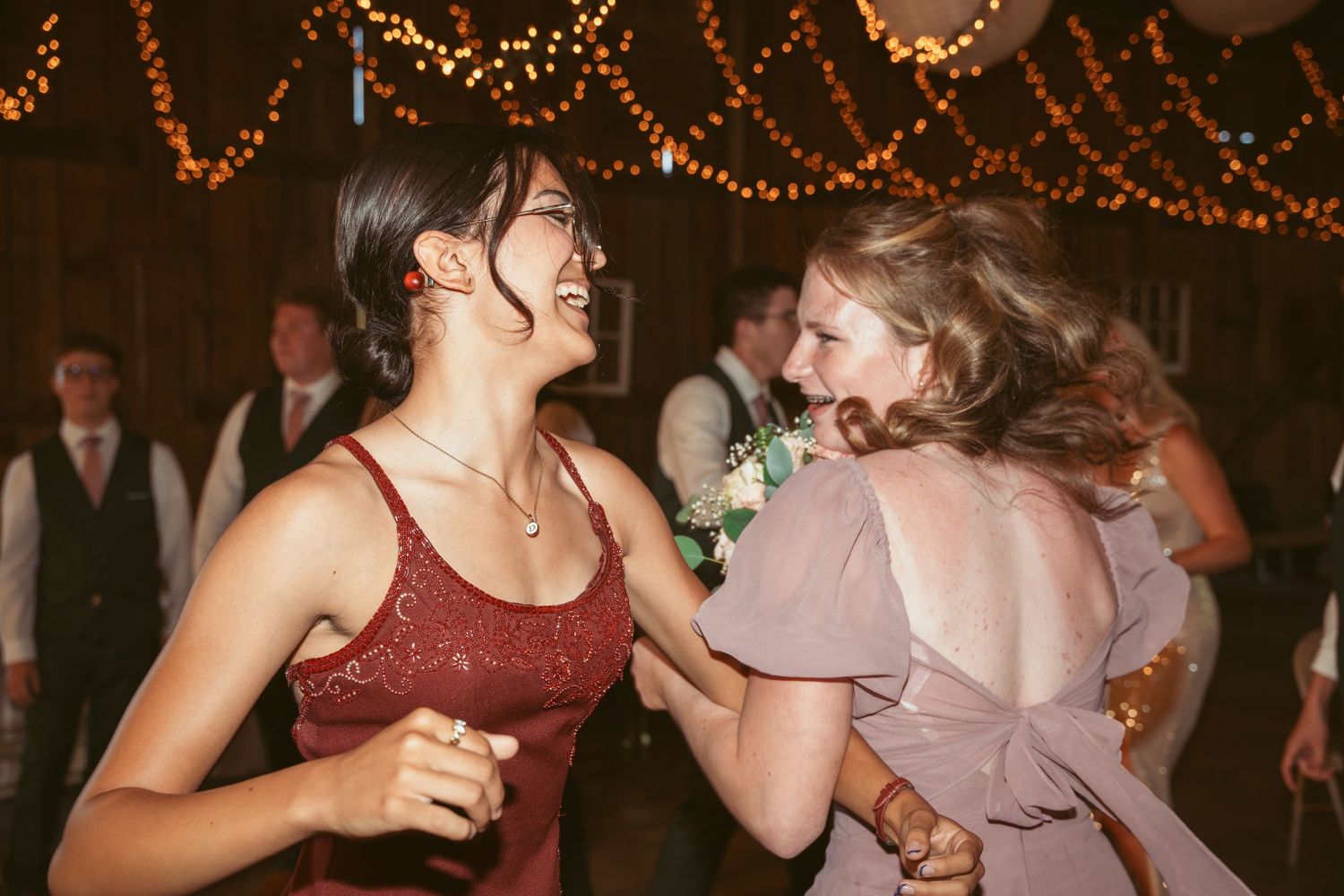 Two guests dance joyfully together under string lights at an evening wedding reception.