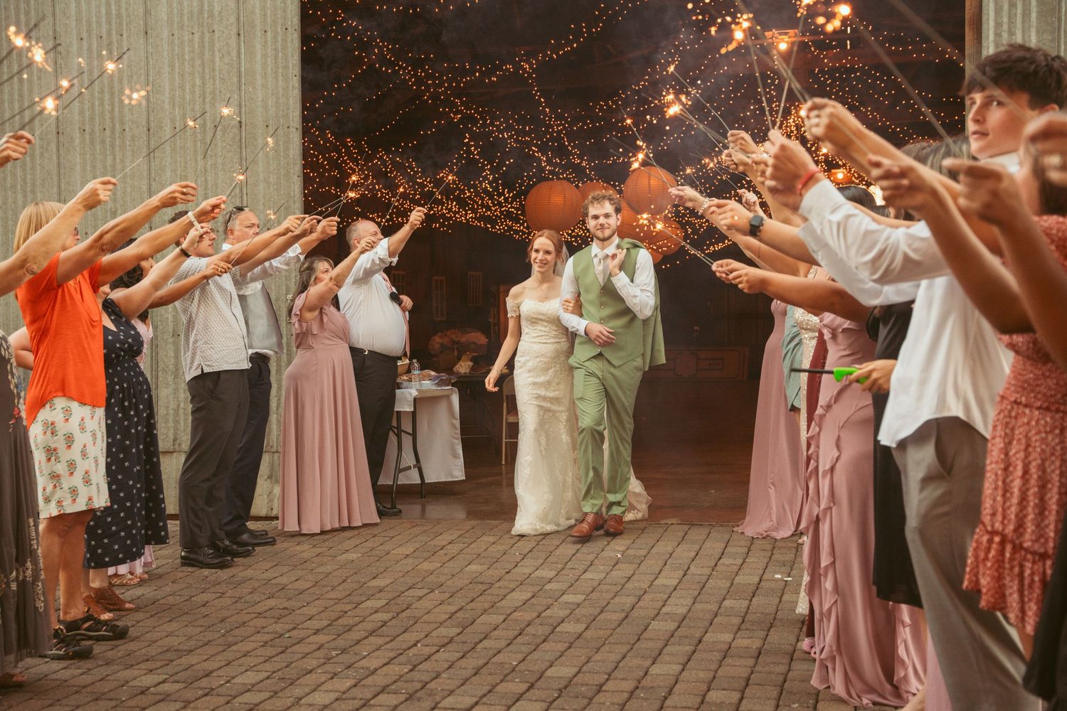 Wedding guests form a sparkler tunnel at dusk for the newlyweds walking through in formal attire.