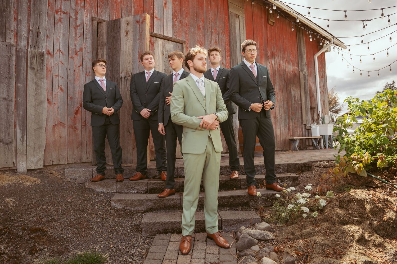 Group photo of wedding party in formal attire against rustic barn backdrop with string lights.