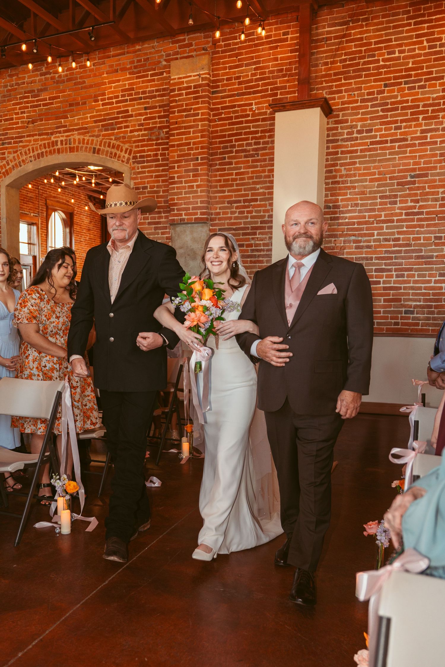 Wedding ceremony guests gather in an industrial venue with exposed brick walls and string lights.