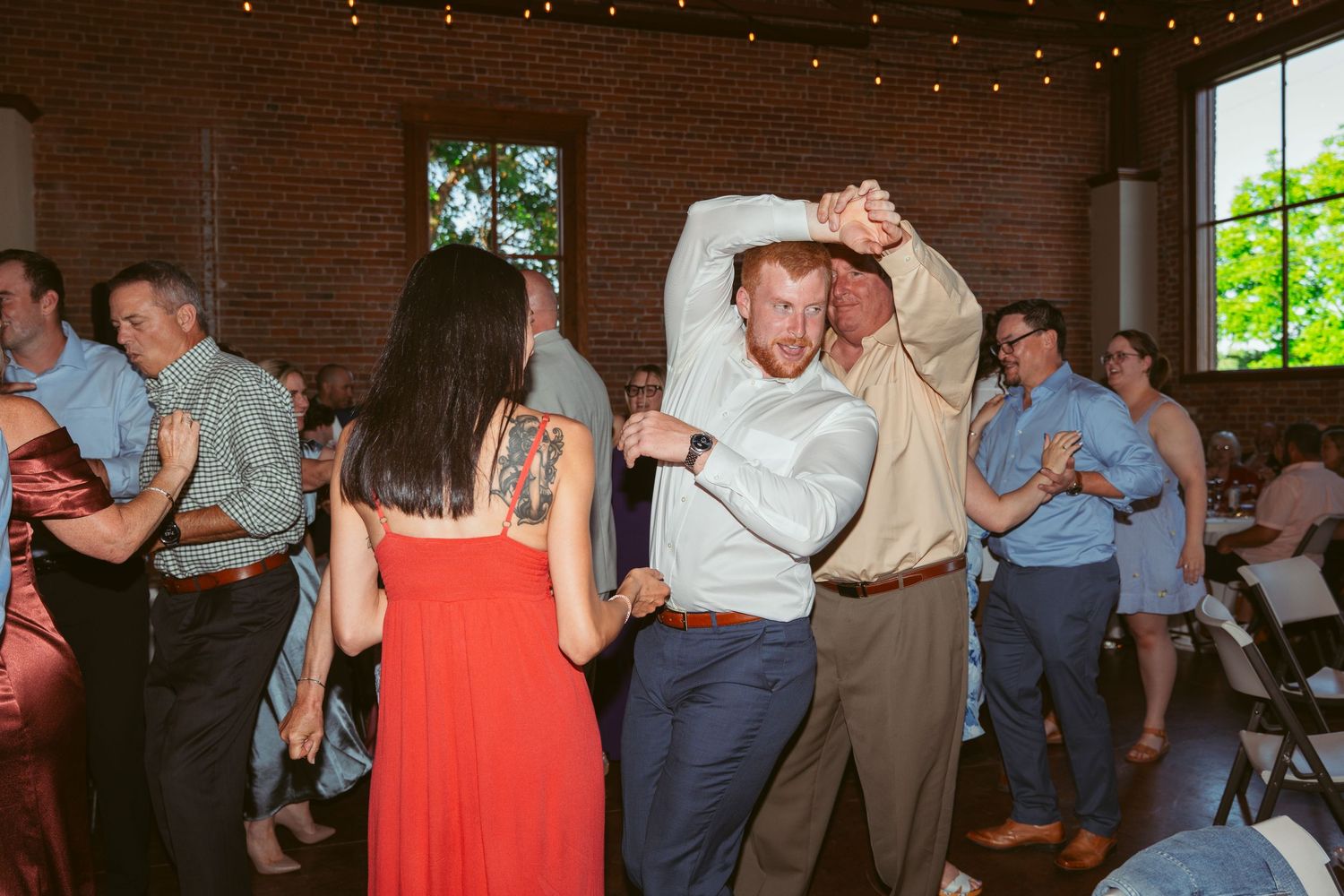 Wedding guests dance and celebrate at a reception in a rustic brick venue with string lights overhead.