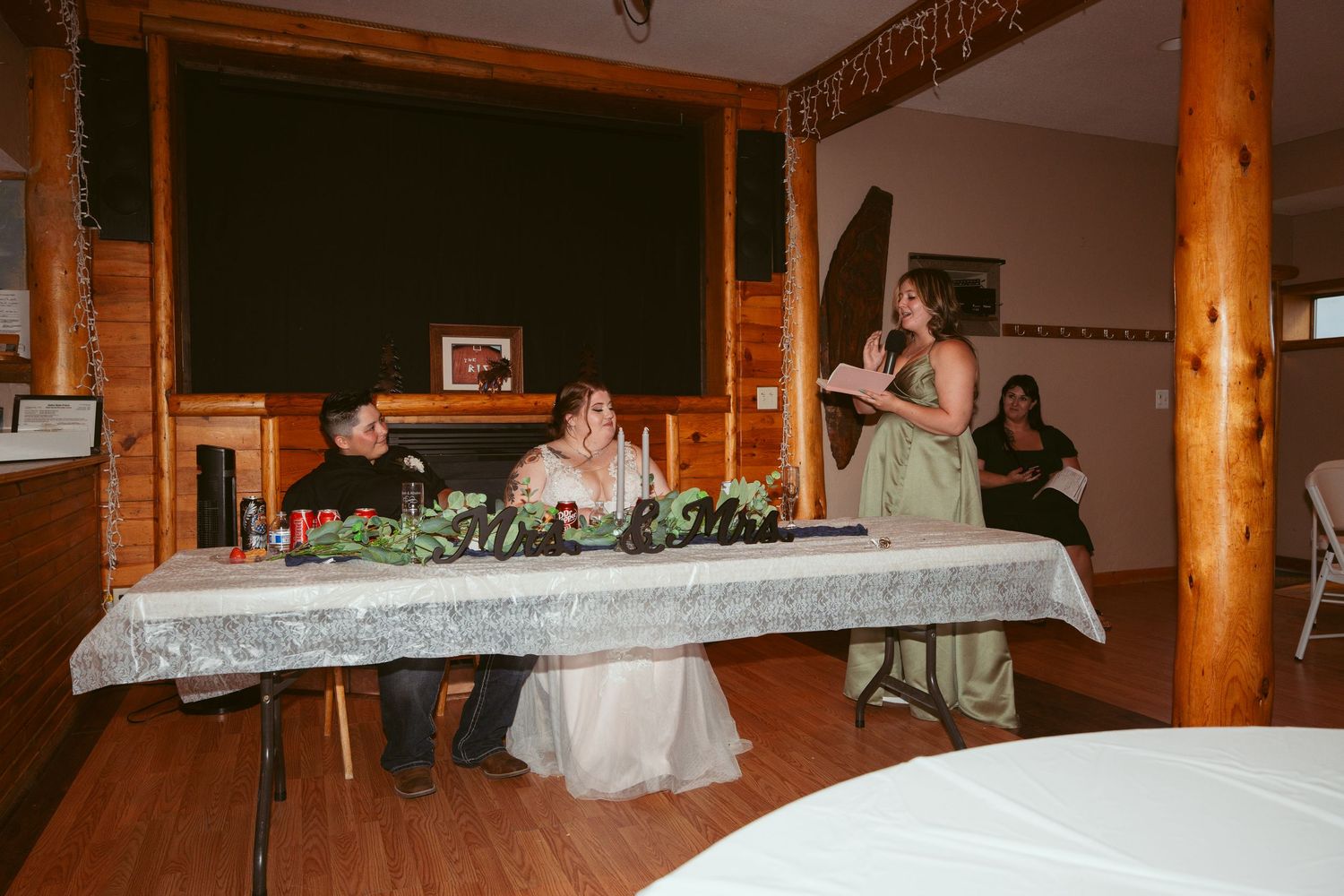 Wedding party seated at decorated head table in rustic venue with wooden beam details.