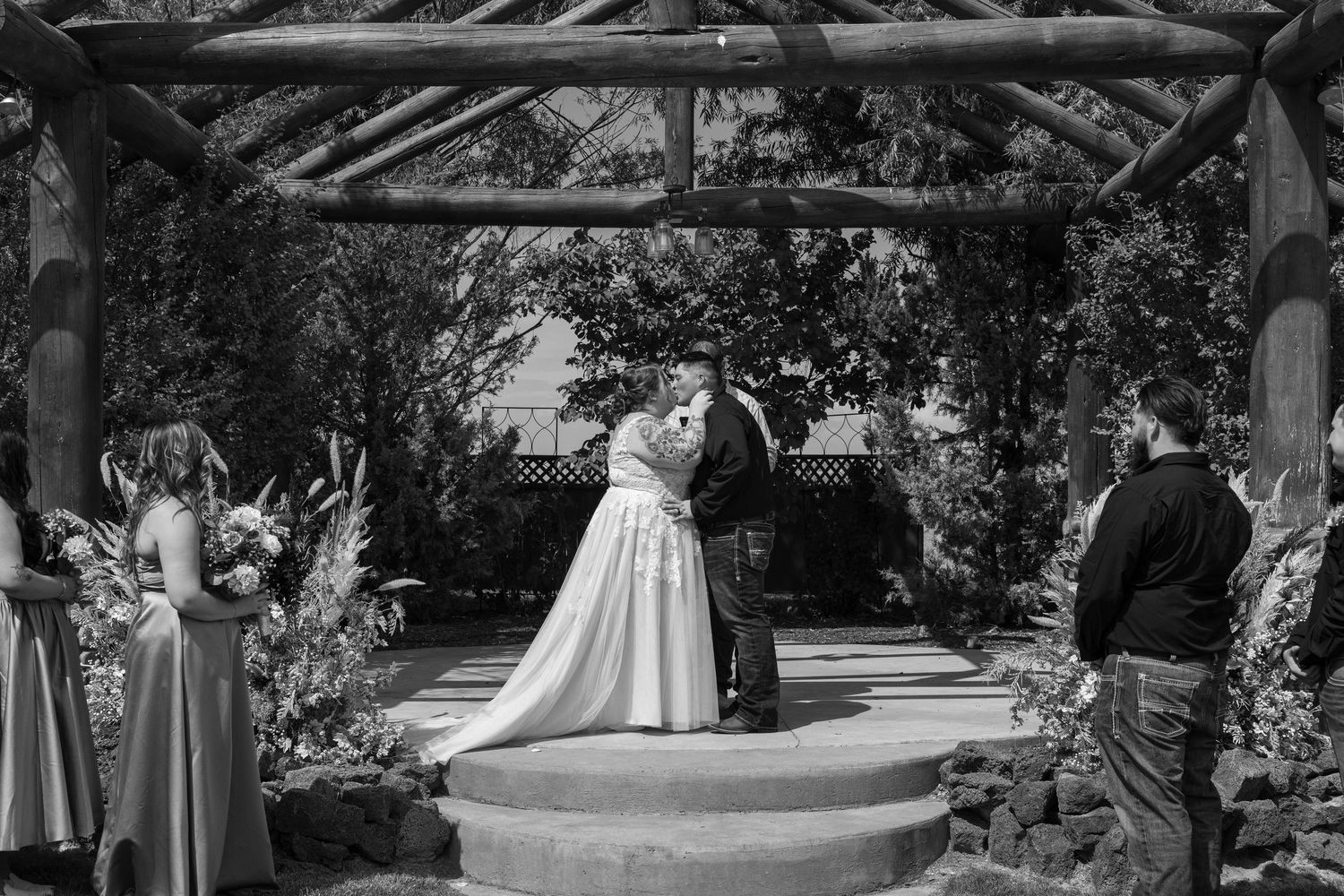 Black and white panoramic photo of a wedding ceremony under a rustic wooden pergola surrounded by trees.