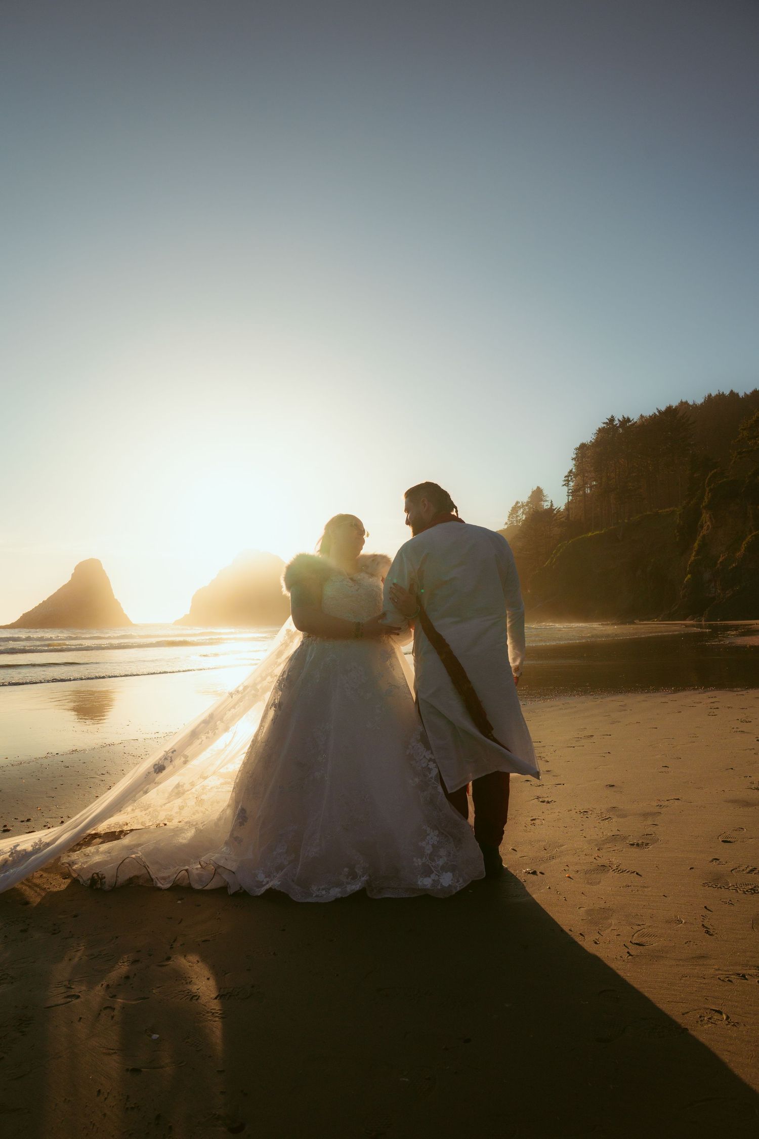 A couple in wedding attire sits together on a beach at sunset with waves and rocky cliffs in view.