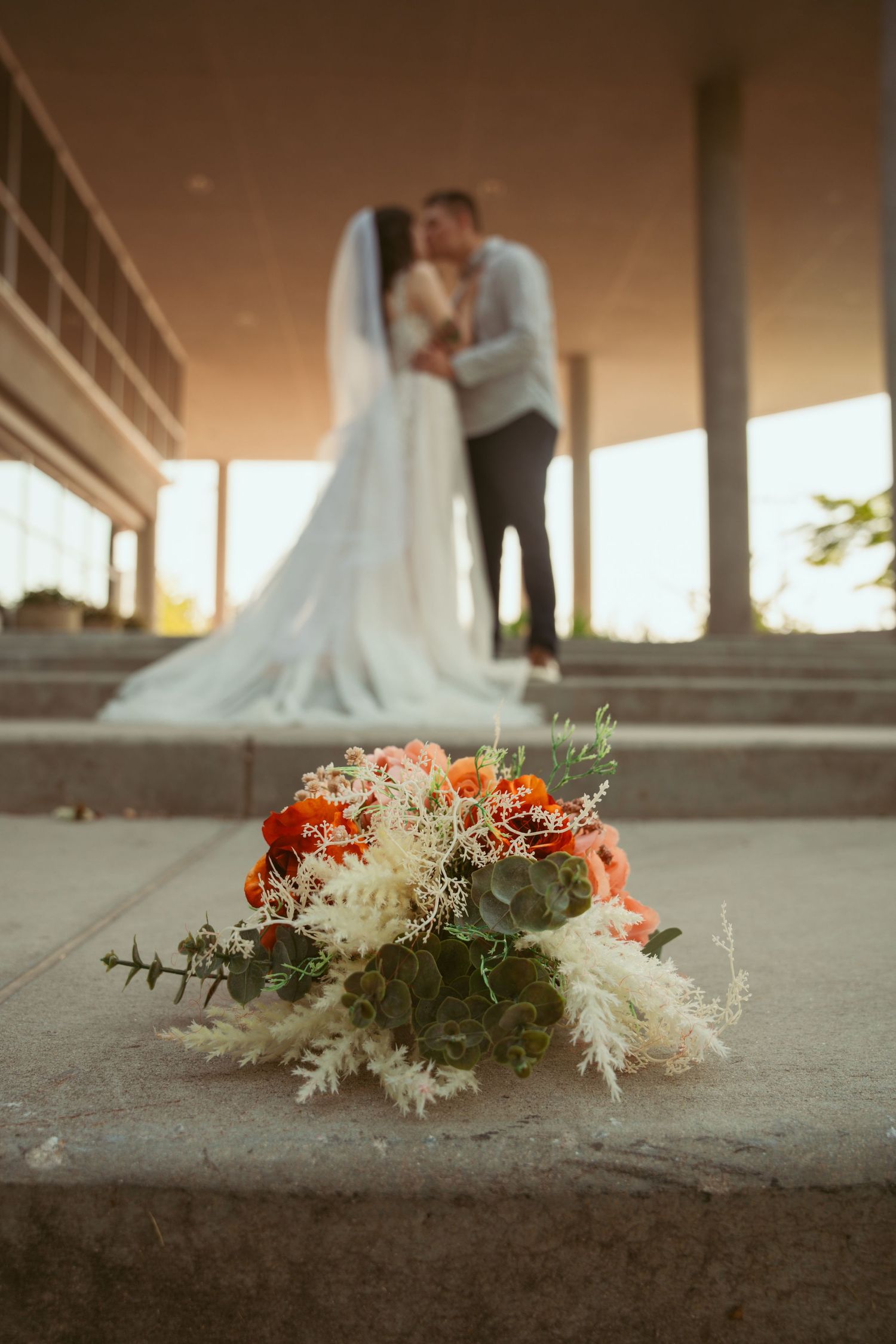 Wedding floral arrangement with white and orange flowers in foreground with couple silhouetted in background.