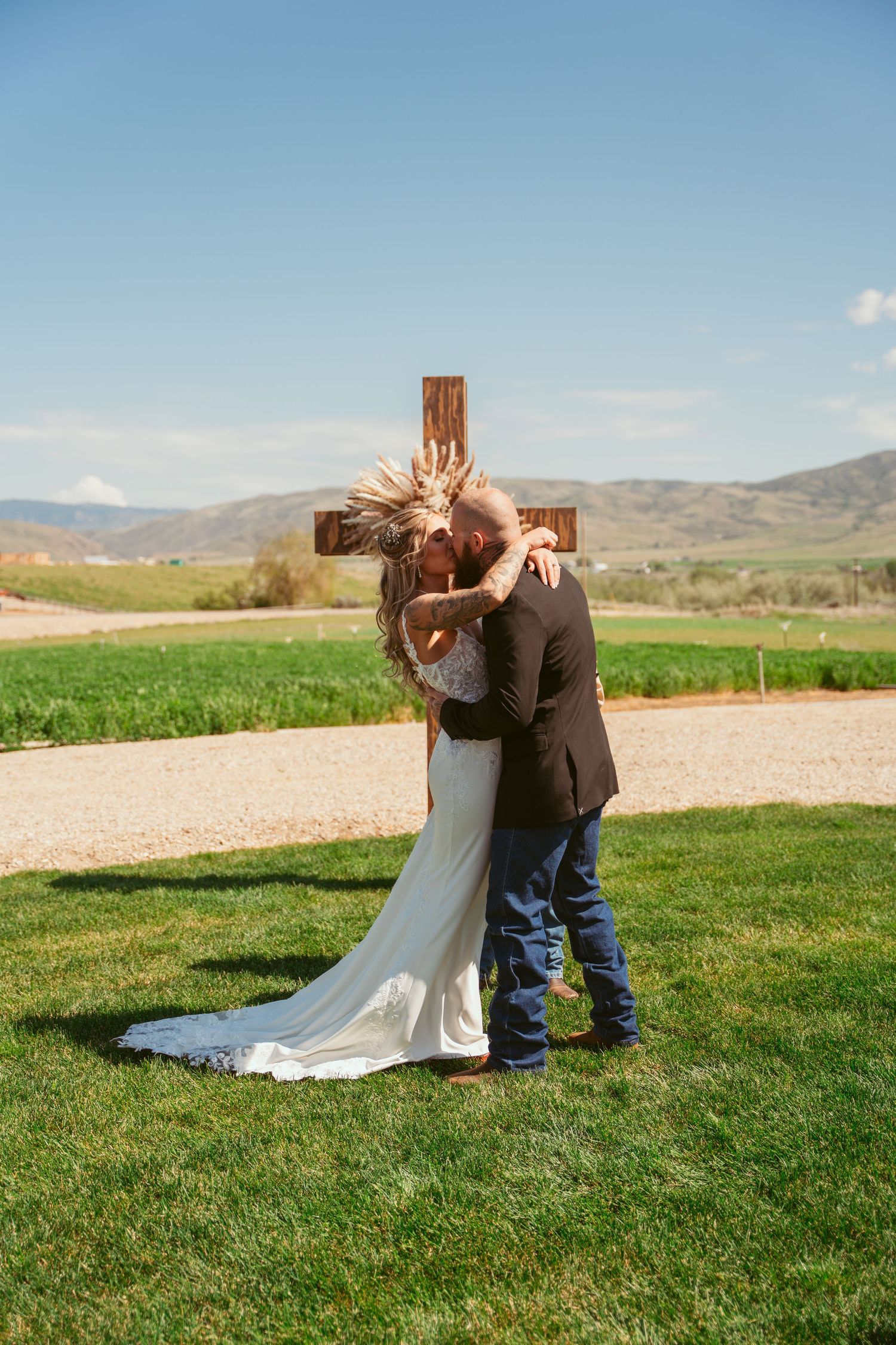 A romantic wedding ceremony moment captured in front of a wooden cross with mountains in the distance.