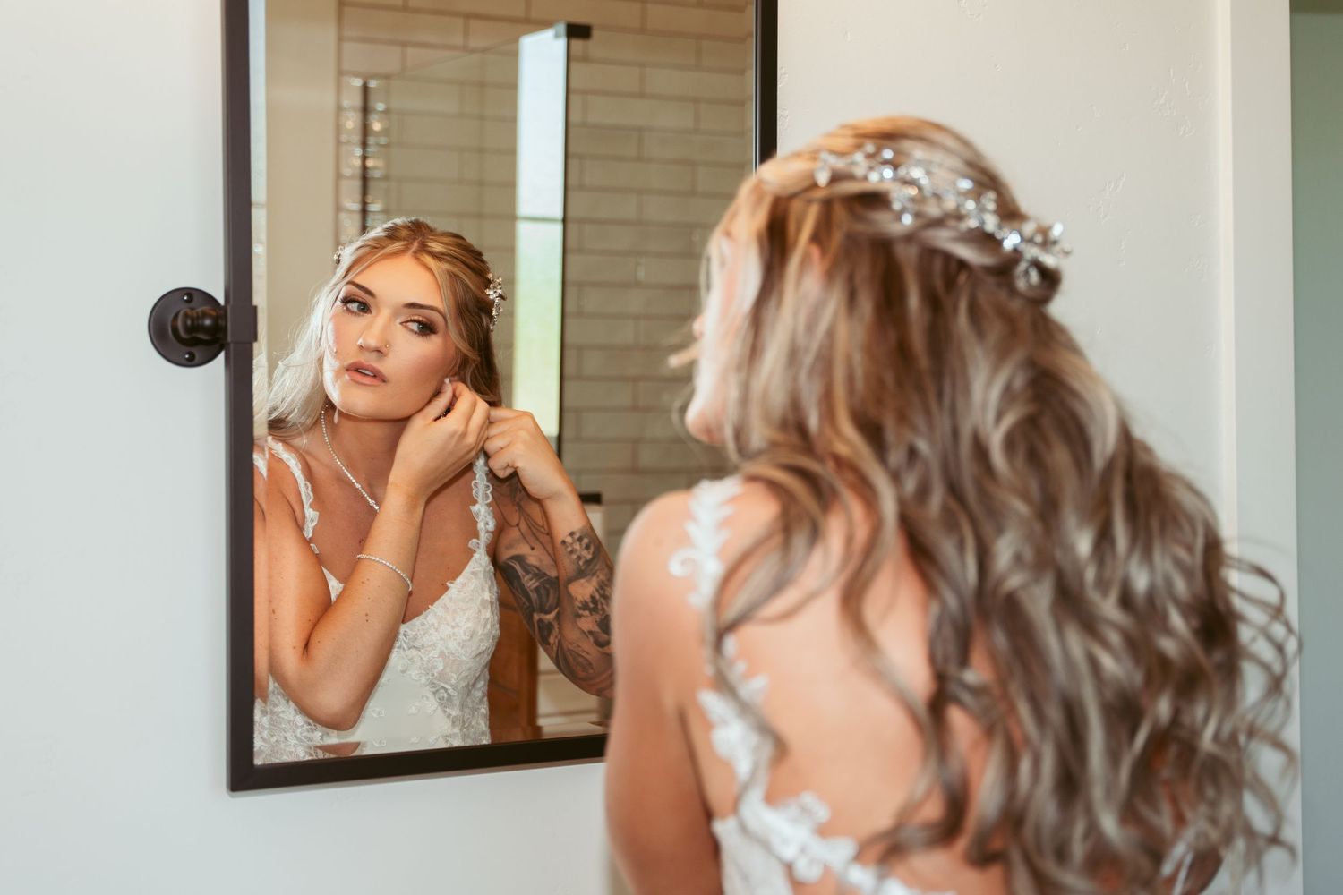 Bride in lace dress putting on earrings while looking in mirror during wedding preparations.