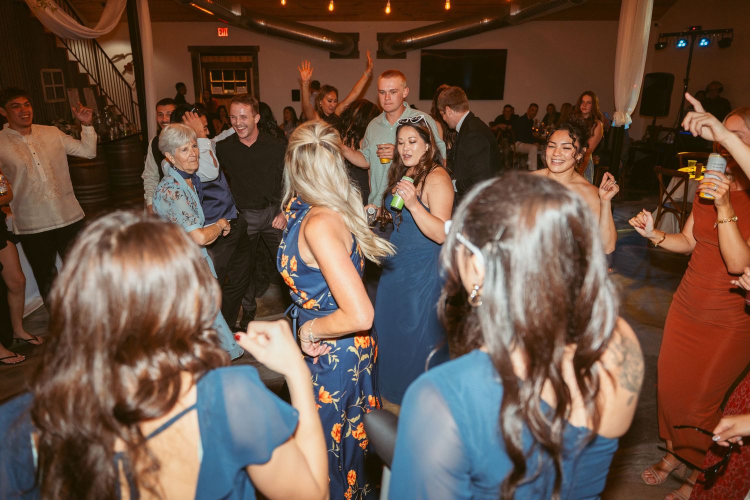 Partygoers in blue dresses dance together under warm string lights at indoor venue.