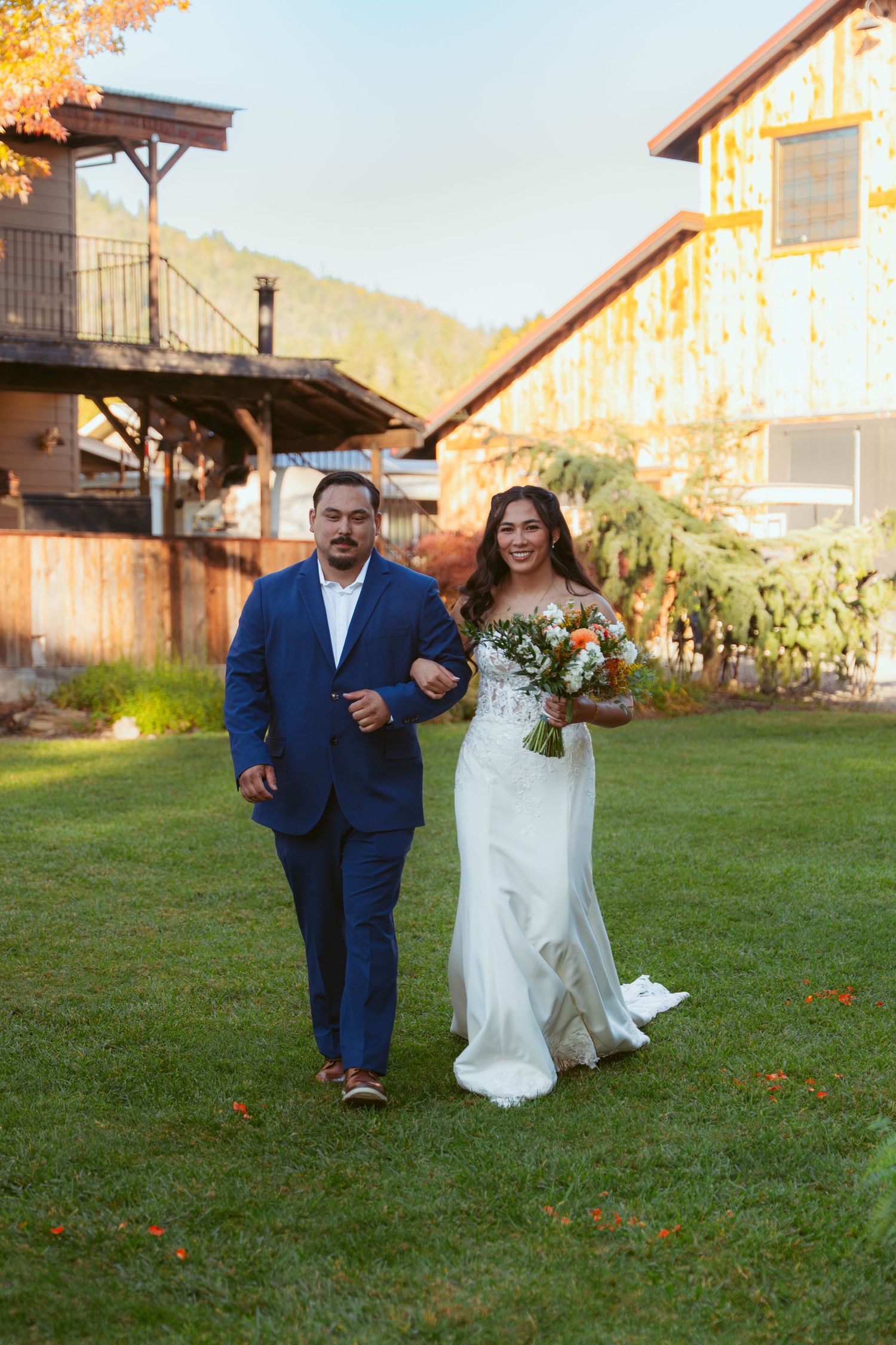 Wedding couple walking across a lawn with rustic barn buildings in the background.