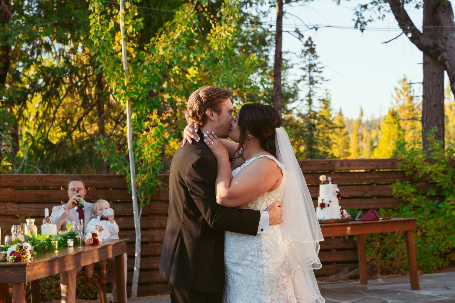 Newlyweds share an intimate dance moment on a stone terrace with wooden fencing and trees in background.
