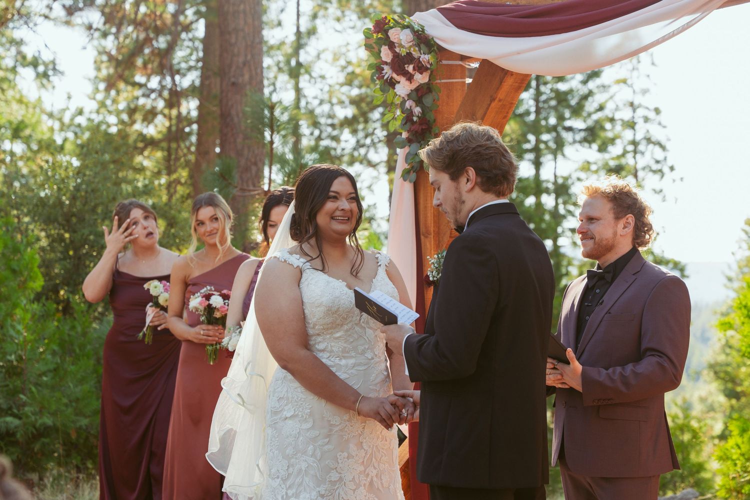 Joyful moments during forest wedding ceremony under wooden arch with flowing fabric.
