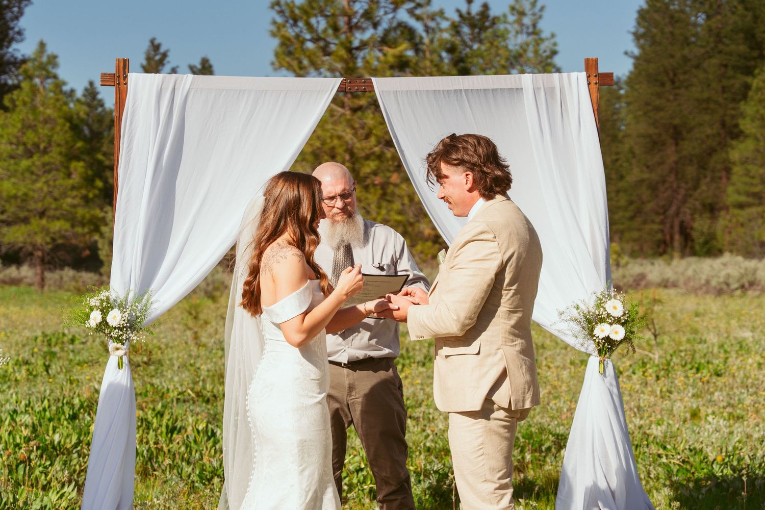Wedding ceremony takes place under white fabric archway in sunny mountain meadow.