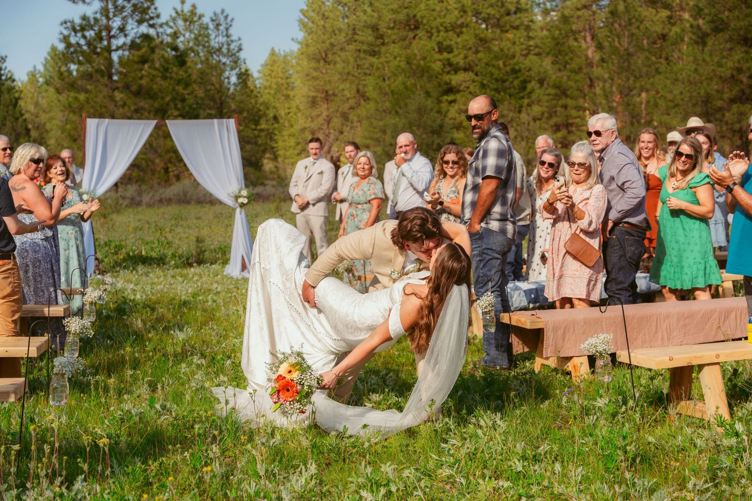 Wedding ceremony in outdoor meadow setting with wooden benches and white arch.