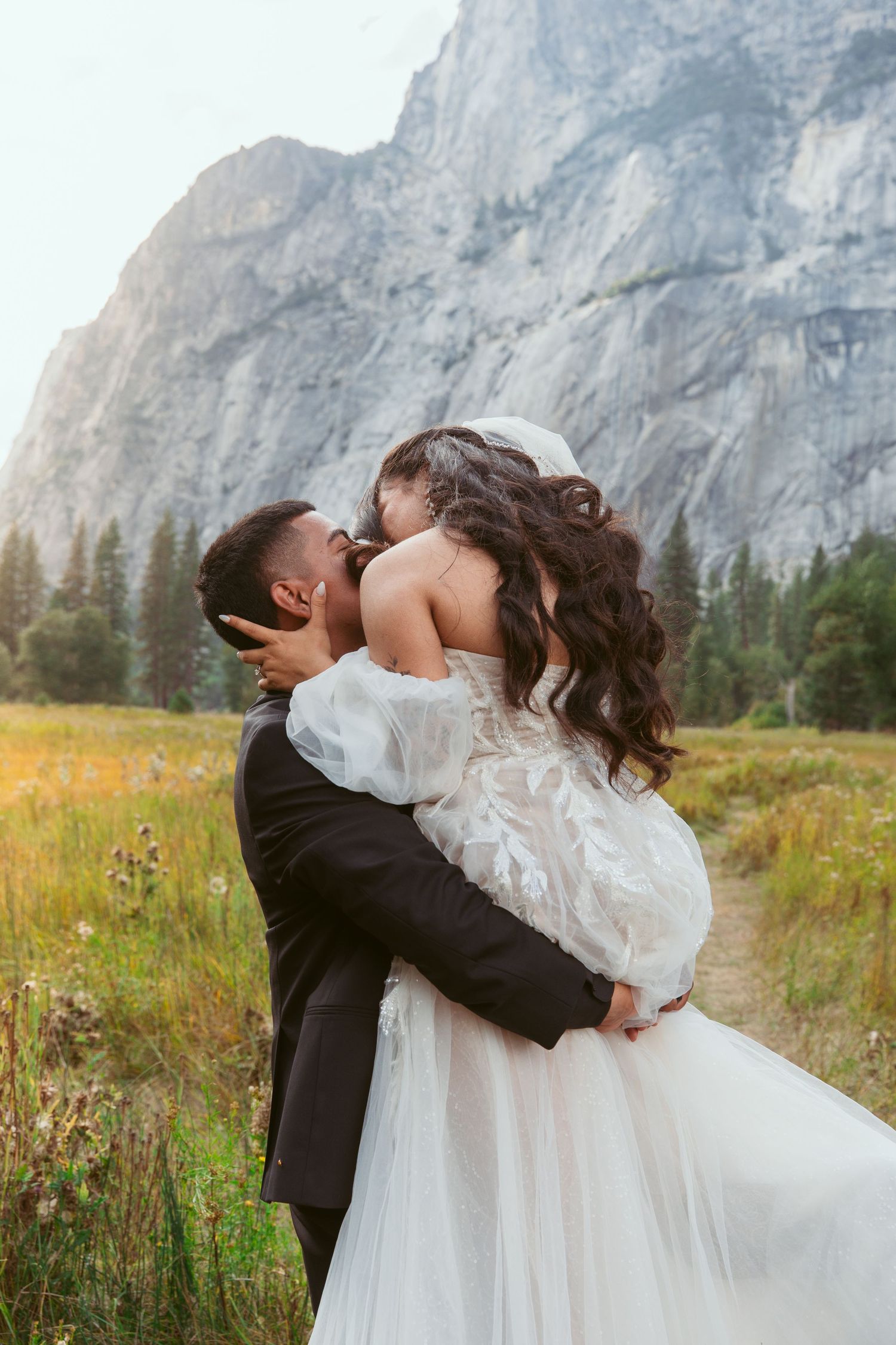 Romantic dip kiss between newlyweds in a mountain meadow with dramatic cliffs in the background.
