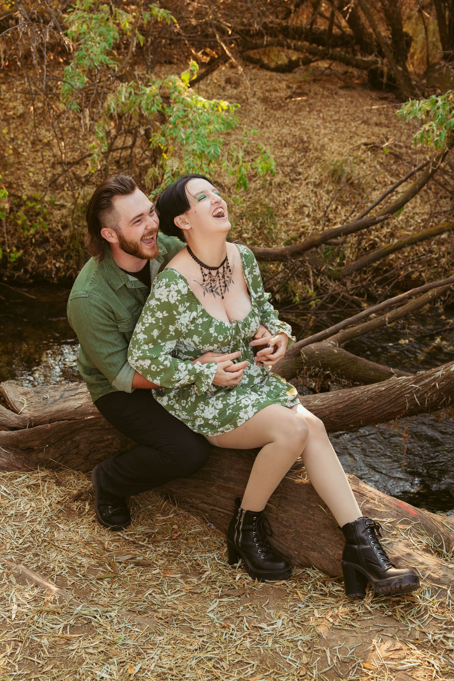 A couple sits together on a fallen log in a wooded area during an autumn photoshoot.