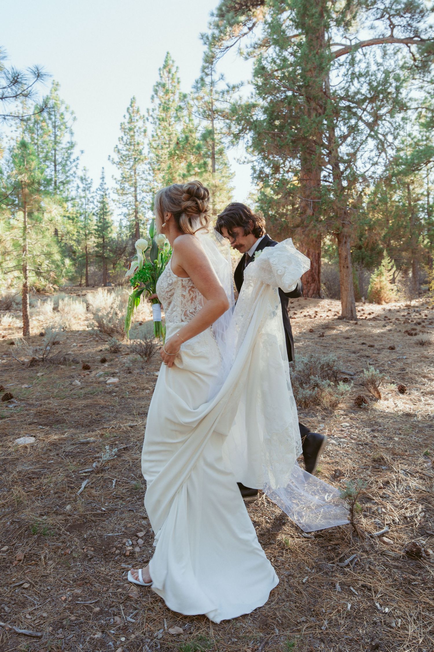 A couple in wedding attire dances playfully on pine needles in a forest setting.