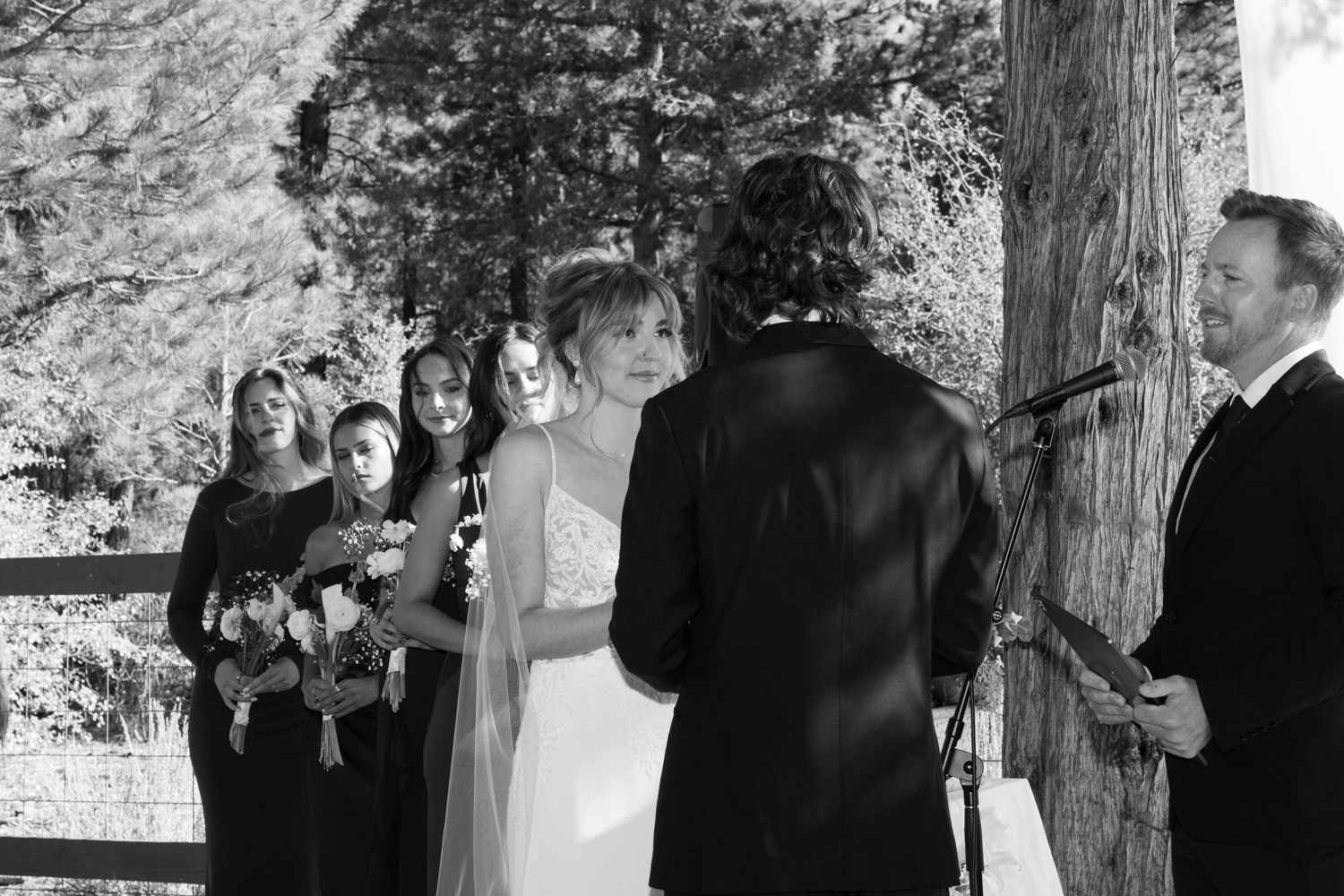 Black and white capture of wedding ceremony with attendants and white backdrop in forest setting.