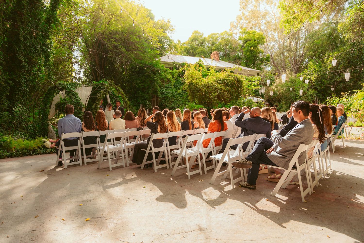Wide angle view of wedding guests seated in white chairs at an outdoor garden venue.