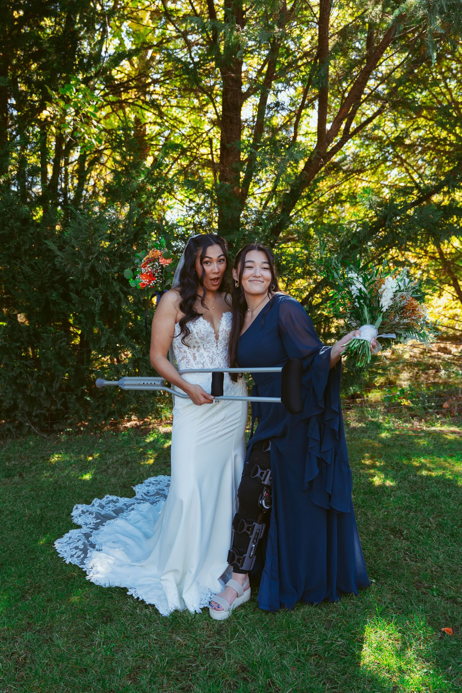 Wedding party members in navy dresses and white gown pose playfully on grassy hill with sunlit trees in background.