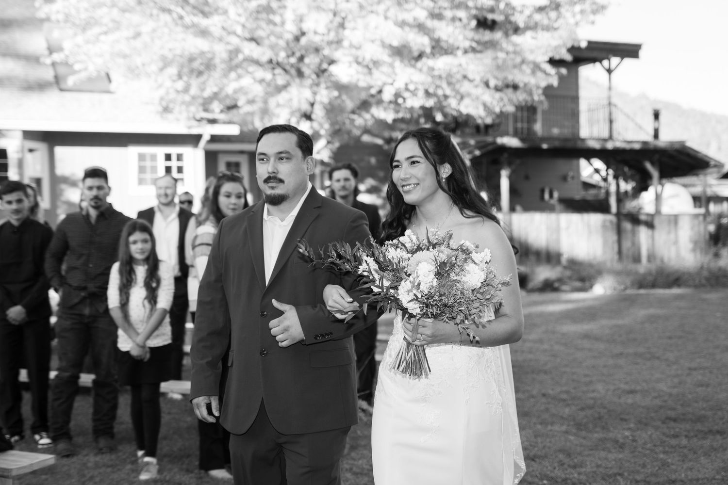 Black and white photo of wedding processional on grass with barn in background.