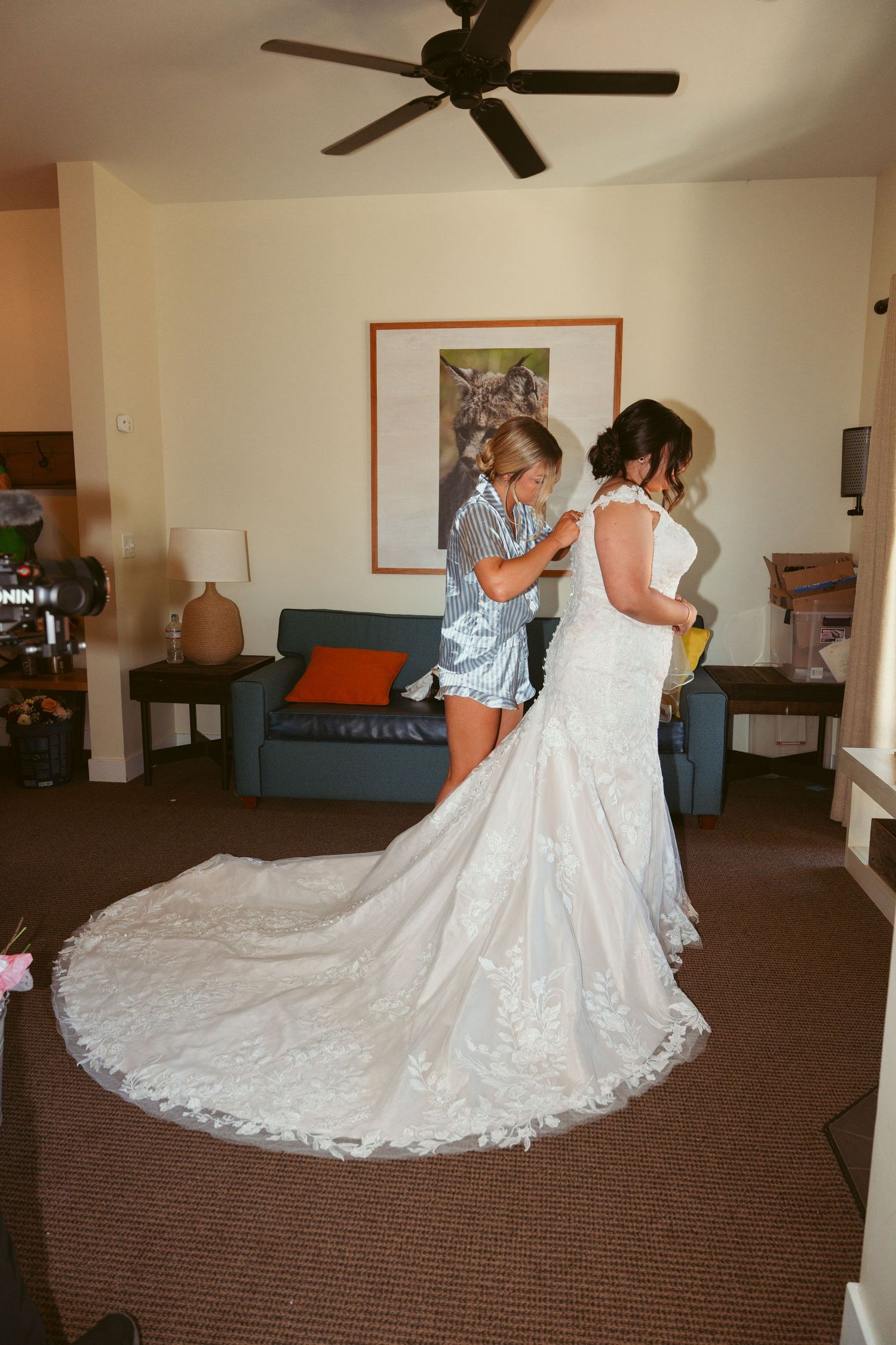 A stunning wedding dress with long lace train being adjusted in a room with ceiling fan.