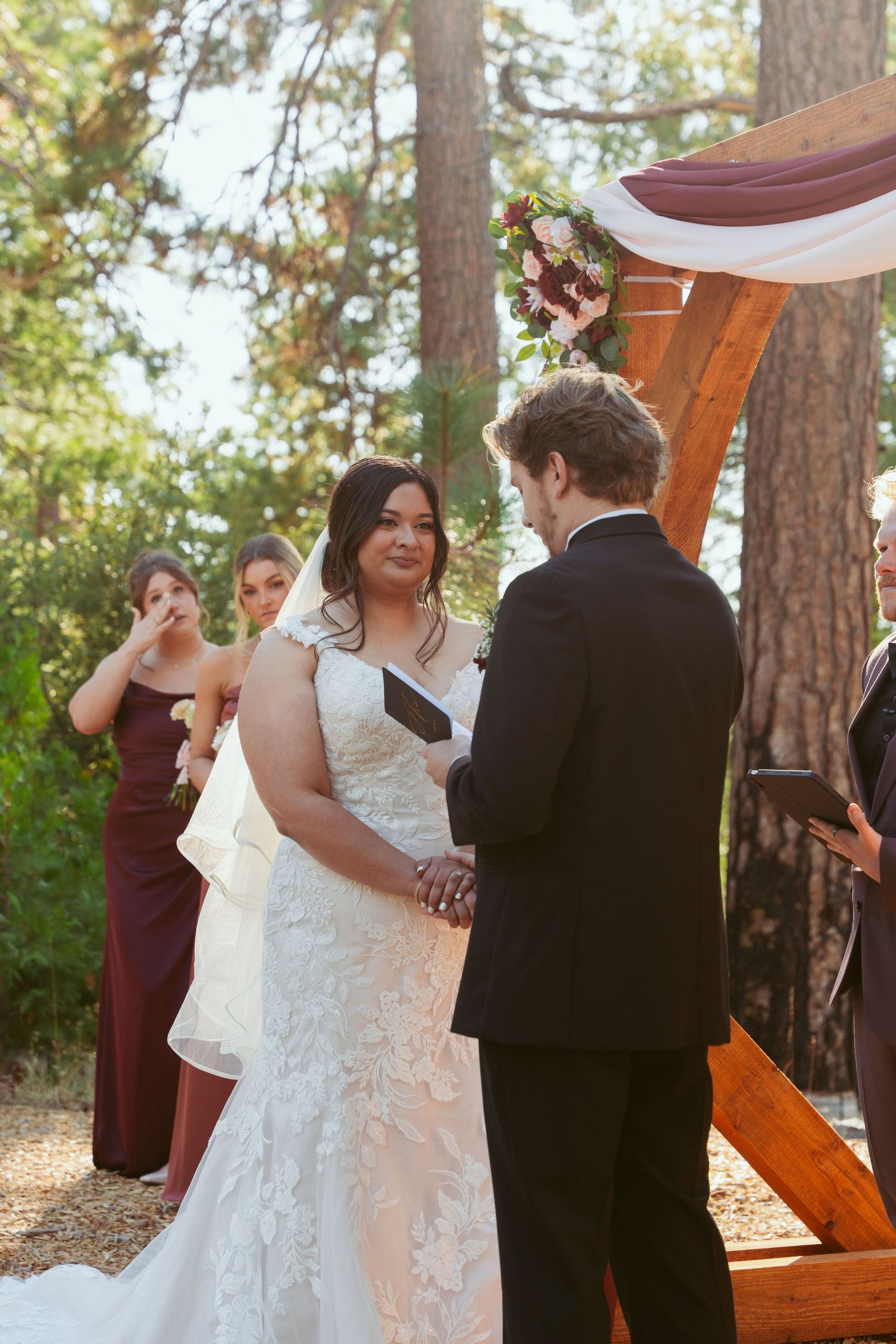 Couple exchanging vows during outdoor wedding ceremony under wooden arch with white draping.