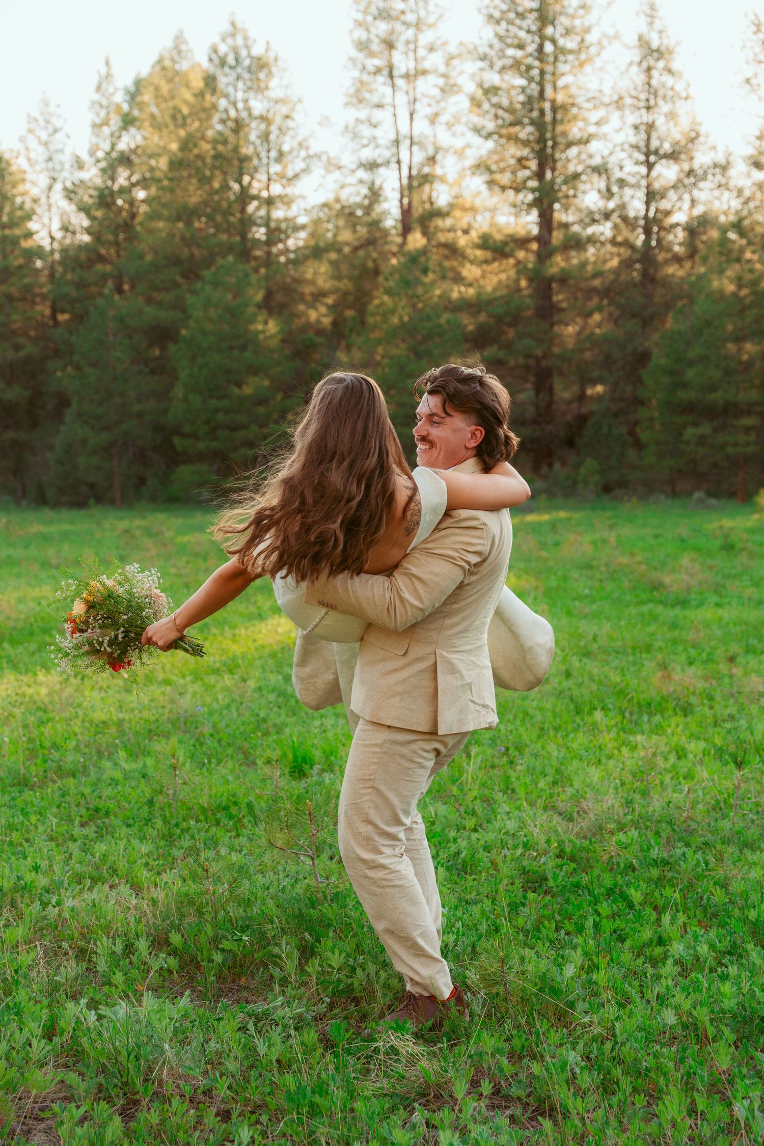 Playful couple dancing and twirling in a green meadow surrounded by evergreen trees.