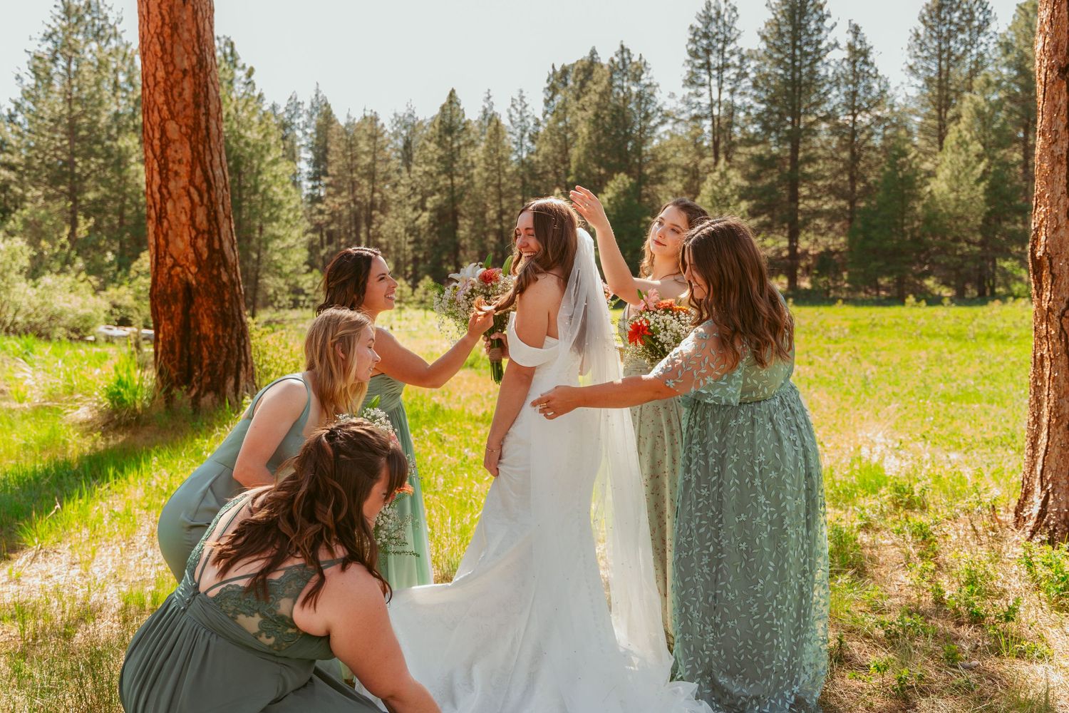 Bridesmaids in flowing green dresses gather outdoors holding bright floral arrangements.