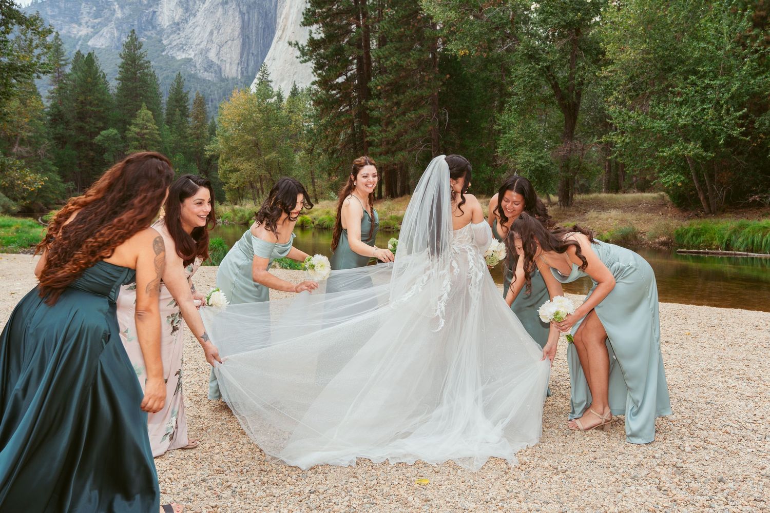 Bridesmaids in blue dresses help arrange bride's dress on scenic mountain beach.