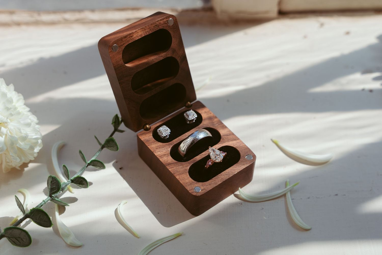 Wooden ring box with wedding bands nestled inside, surrounded by delicate eucalyptus leaves and white flowers.